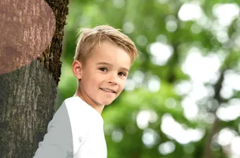 Et smilende barn i hvit skjorte står ved siden av en trestamme